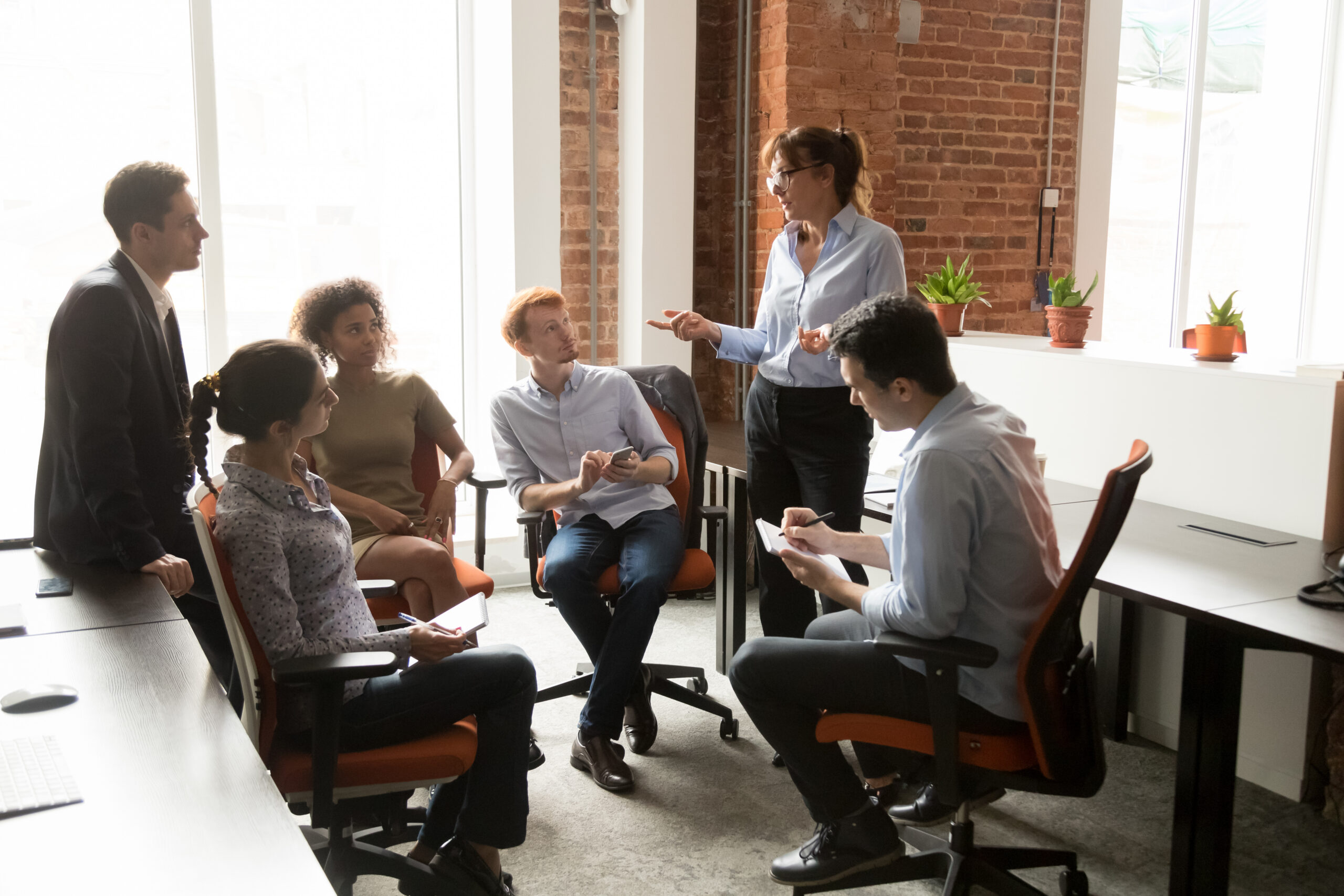 Group of colleagues in a modern office listening to a woman standing and speaking.