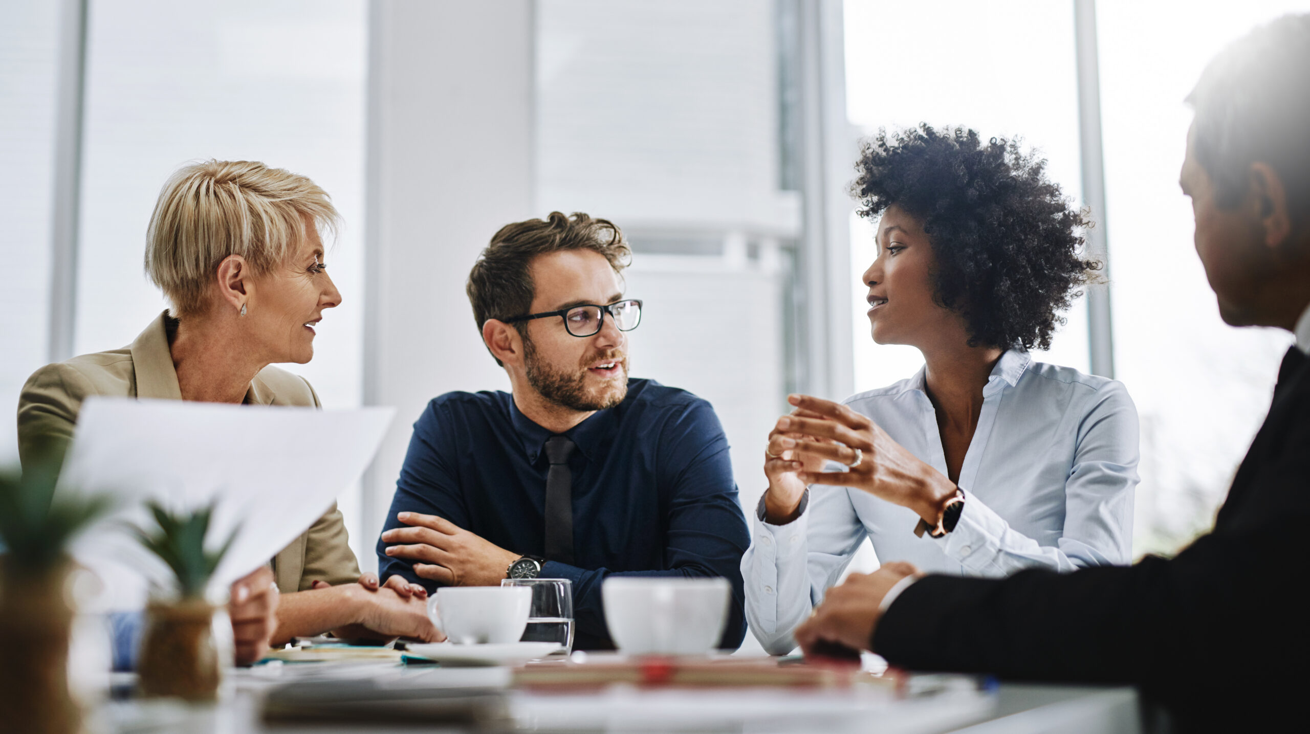Four professionals in a bright office discussing ideas around a conference table with papers and cups ready for coffee.