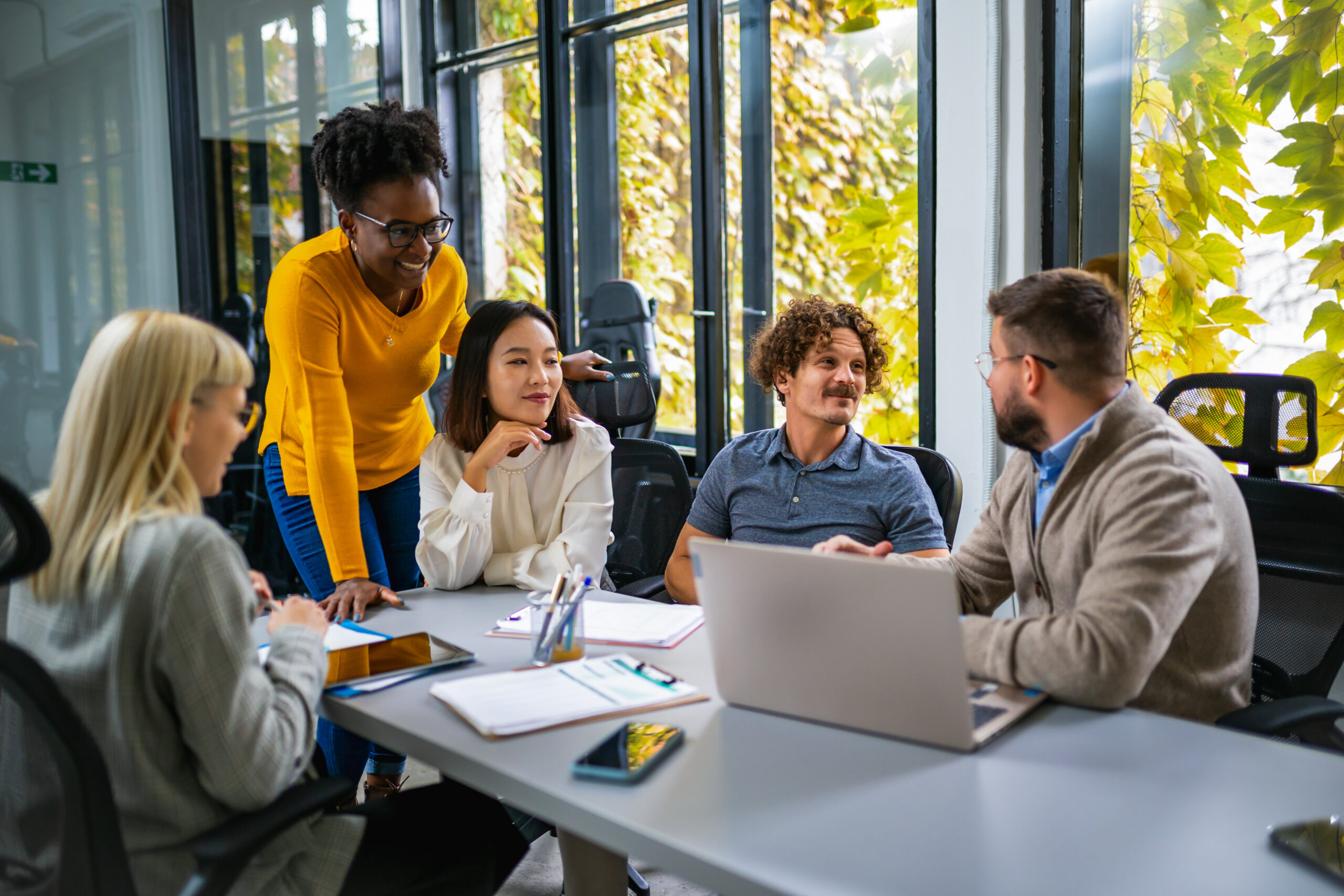 Coworkers in a bright office around a conference table, a woman in a yellow sweater leaning in to talk to teammates.