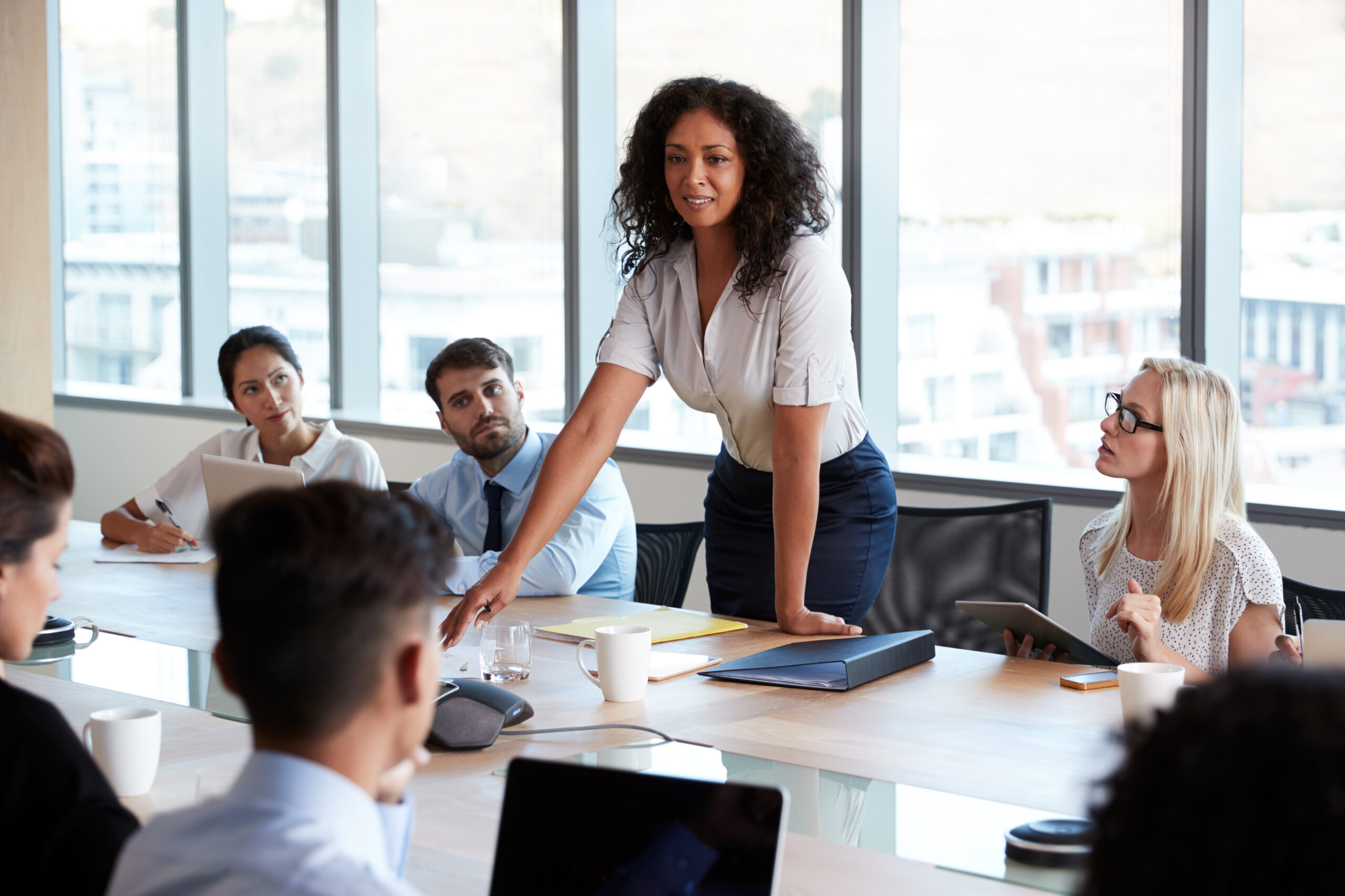 Woman standing and presenting at a conference table as colleagues listen in a bright, modern office.