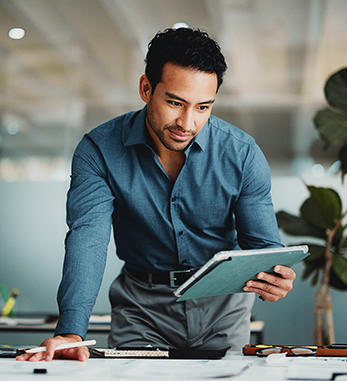Businessman in a blue shirt leaning over a desk, examining documents with a tablet in hand in a modern office.