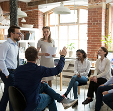 Team of six coworkers in a brick-walled office, with one person presenting papers while others listen.