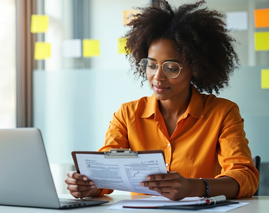 Woman in an orange shirt reviews documents on a clipboard at a desk, with a laptop and sticky notes in the background.