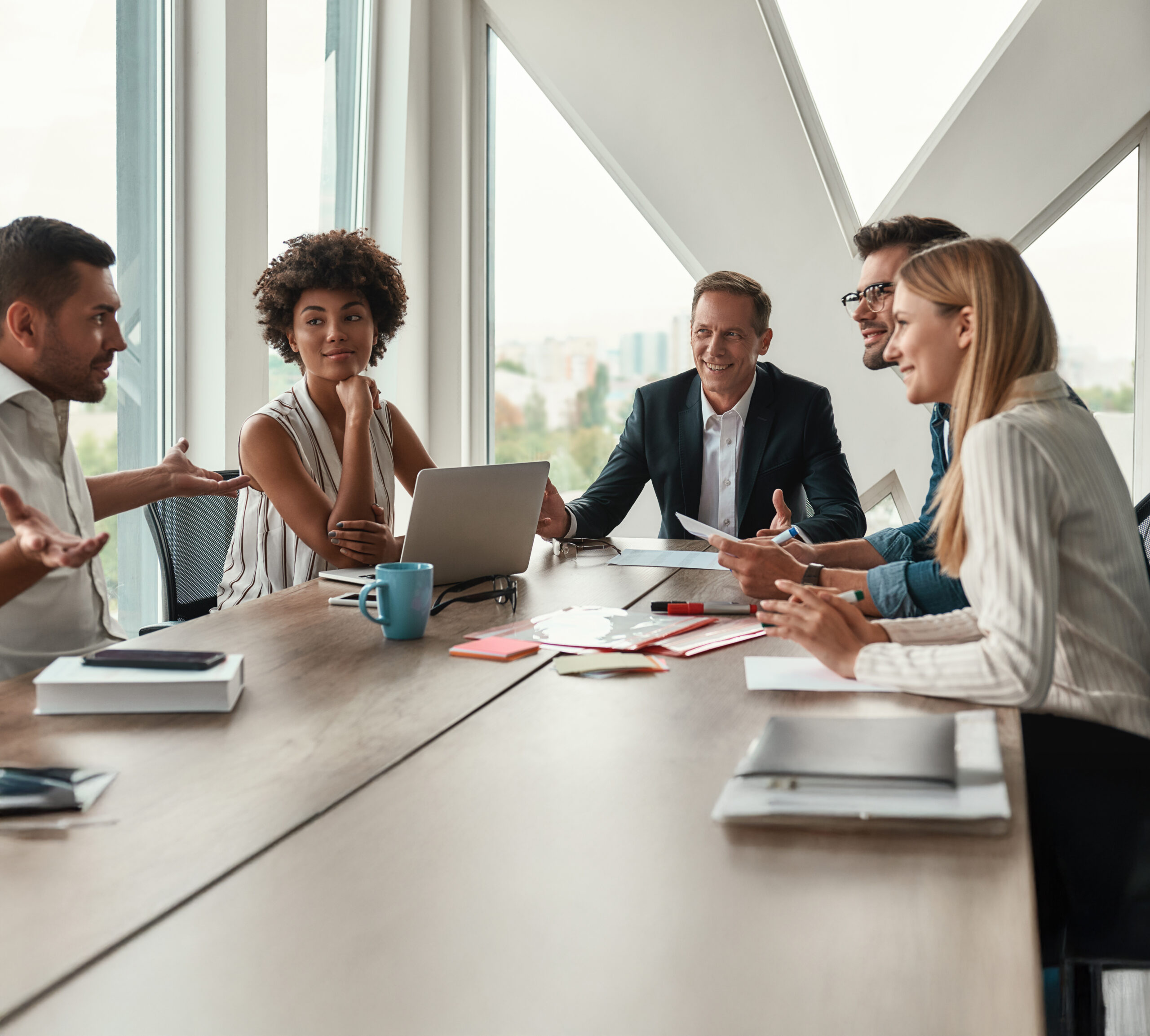 A diverse group of professionals seated around a large conference table, talking and sharing documents by a windowed wall.